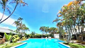 Ein rechteckiger Swimmingpool mit klar blauem Wasser, umgeben von gepflegtem Rasen und verschiedenen tropischen Pflanzen.