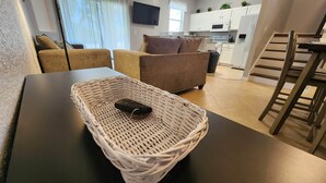 Een woonkamer met een donkere tafel, een rieten mand met een afstandsbediening, een bruine bank en een keuken op de achtergrond.