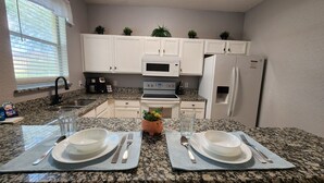 Een keuken met witte kastjes, een magnetron, kookplaat, koelkast, granieten aanrechtbladen en twee eettafels gedekt met borden, bestek en glazen.