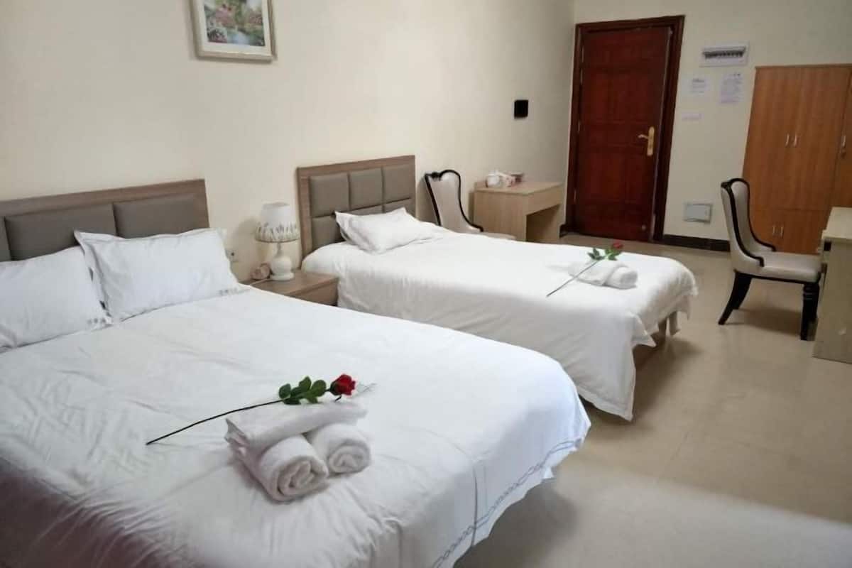 Habitación de hotel con dos camas, cada una con ropa blanca y una sola rosa roja. Hay una silla y una mesita con una lámpara.
