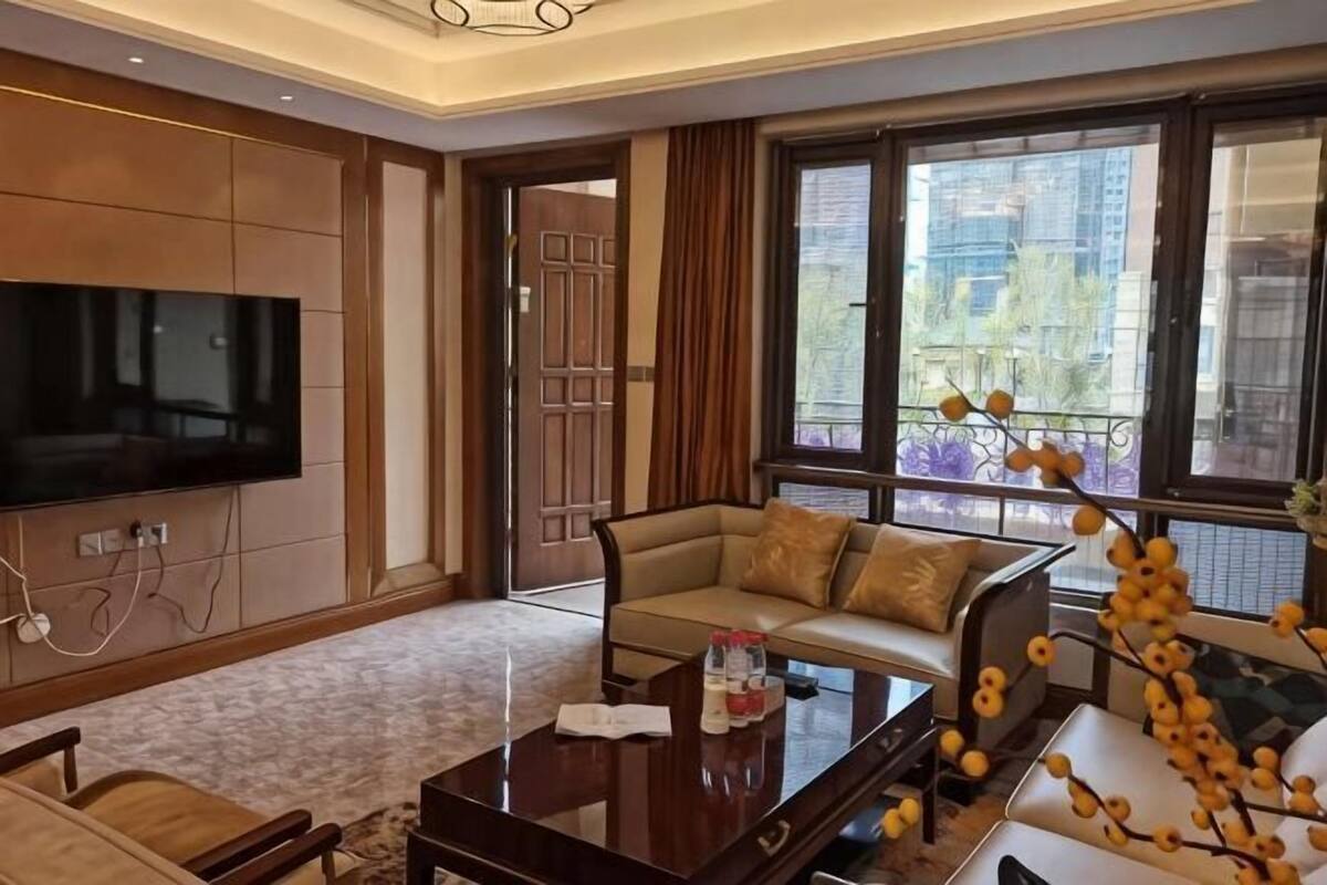 Una sala de estar moderna con televisor de pantalla plana, sofá, mesa de centro y ventanas amplias que ofrecen vistas a edificios y árboles.