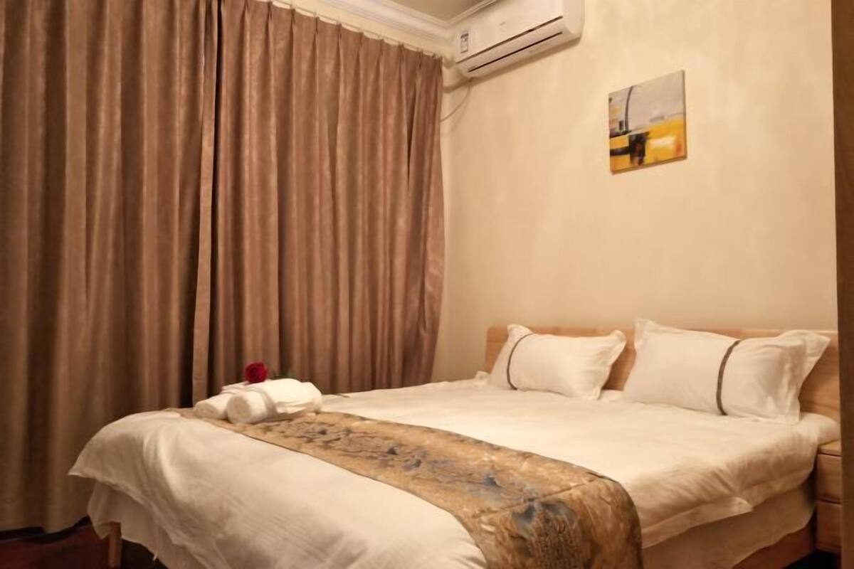 Una habitación de hotel con cama, cortinas color beige, un equipo de aire acondicionado y un cuadro en la pared.