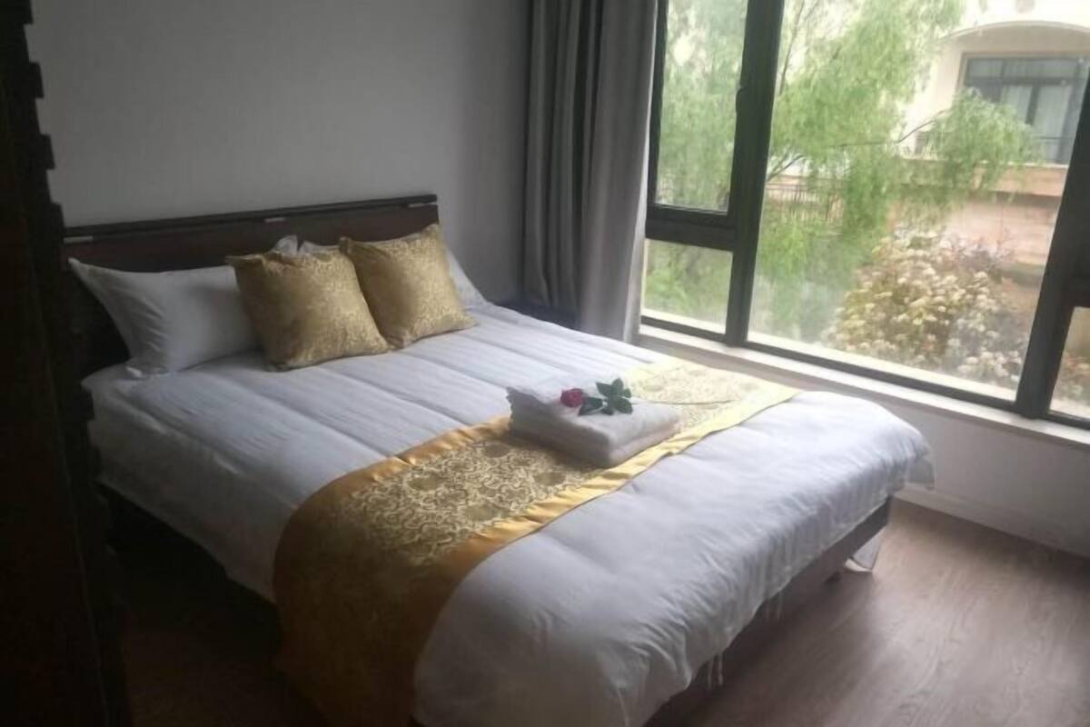 Una cama con ropa de cama blanca, un detalle dorado y una ventana con vistas a la vegetación.