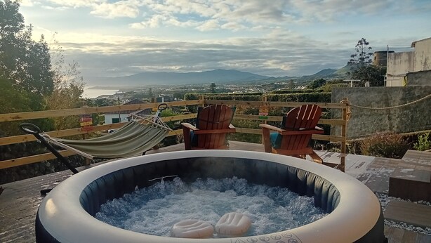 A hot tub with two inflatable rings, a hammock, and wooden chairs on a rooftop with a view of a city and mountains.