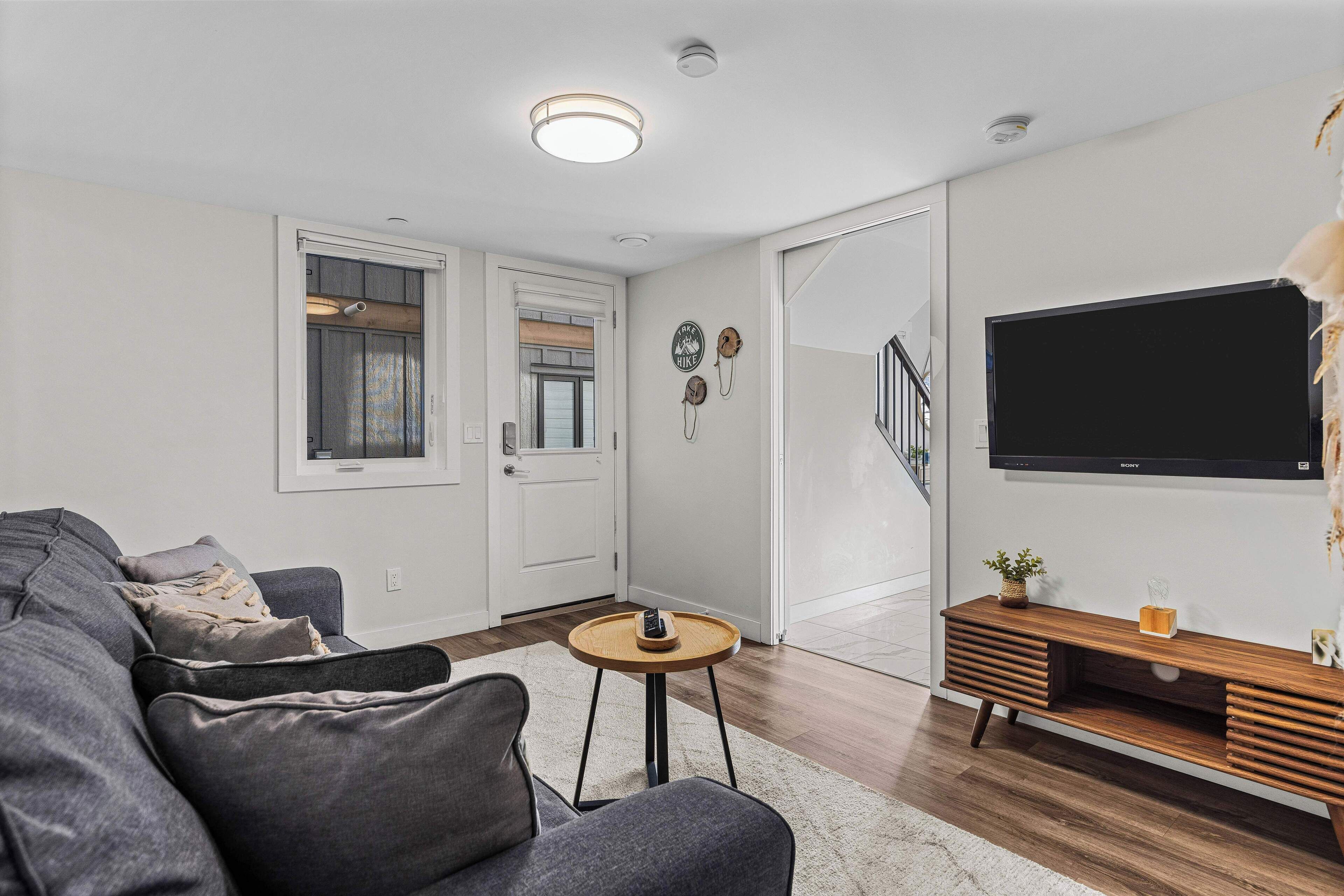 Een moderne woonkamer met een grijze bank, een kleine ronde tafel, een houten tv-meubel en een trap die naar een ander niveau leidt.