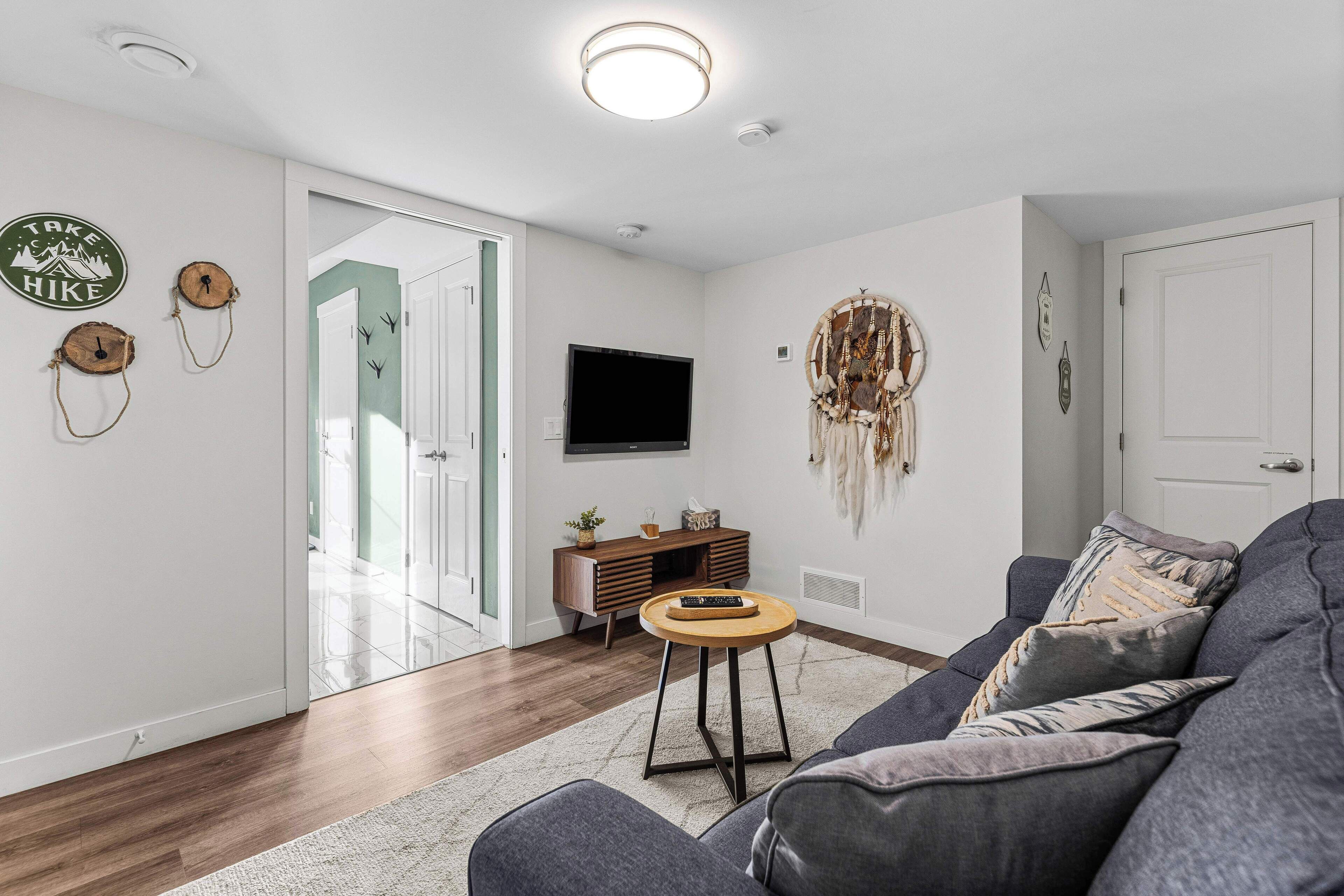 A modern living room with a grey sofa, a round wooden coffee table, a flat-screen TV, and a wall-mounted dreamcatcher.