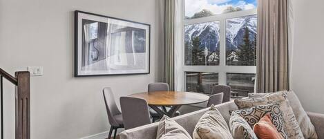 A modern living room with a wooden dining table, grey sofa, and a large window with a mountain view.