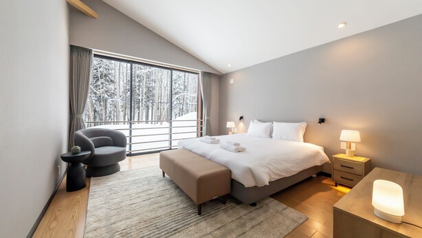 A modern bedroom with a large bed, bedside tables, a bench, and a view of a snowy forest through large windows. - Mori Casa (Kutchan)