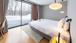 A modern bedroom with a large bed, a bedside table with a lamp, a view of a snowy landscape through a sliding door, and a wooden floor. - Mori Casa (Kutchan)