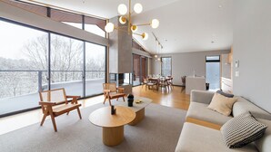 A modern living room with a sofa, coffee table, and chairs. There is a fireplace, large windows, and a dining area in the background. - Mori Casa (Kutchan)