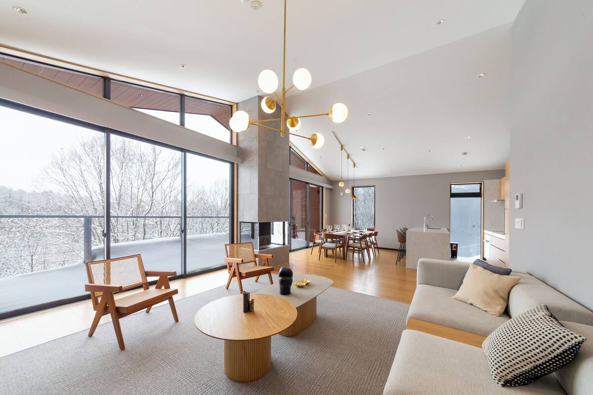 A modern living room with a sofa, coffee table, and chairs. There is a fireplace, large windows, and a dining area in the background.