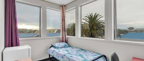 A small, single-bed room with a view of palm trees and buildings through large windows.