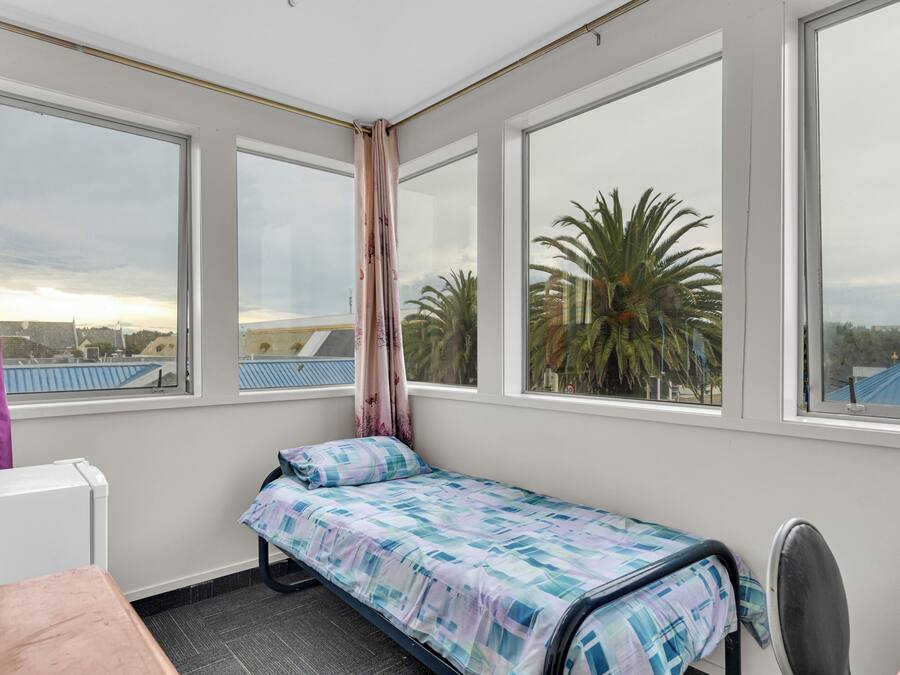 A small, single-bed room with a view of palm trees and buildings through large windows.