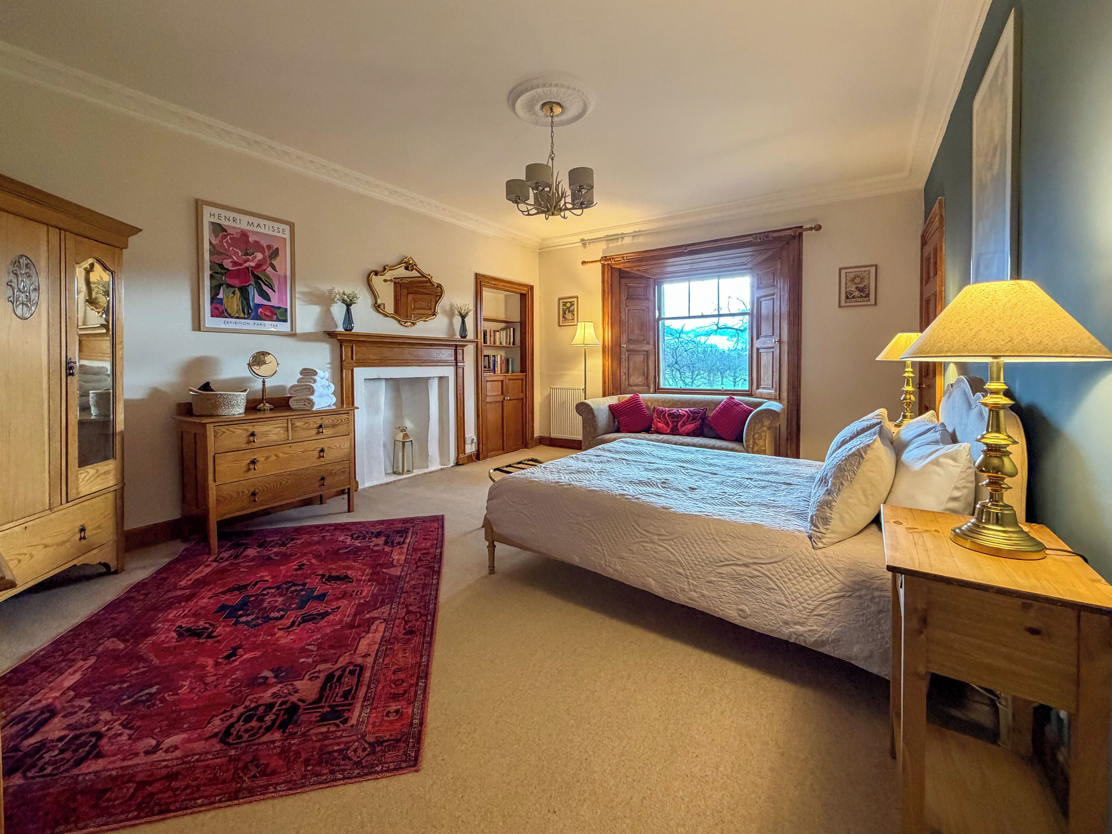 Une chambre à coucher avec un lit, une commode en bois, un miroir, une cheminée, une fenêtre et une lampe.