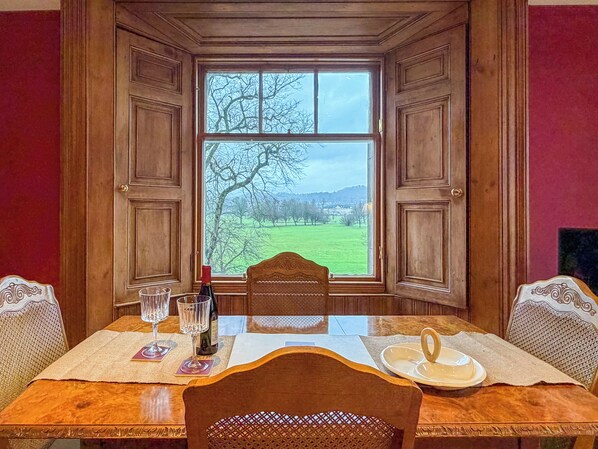 A dining table with a bottle of wine, glasses, and a plate set in a room with wooden paneled walls and a large window overlooking a green landscape.