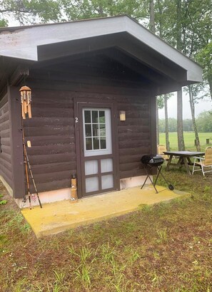 Una cabaña de madera pequeña con porche, una caña de pescar y una parrilla.