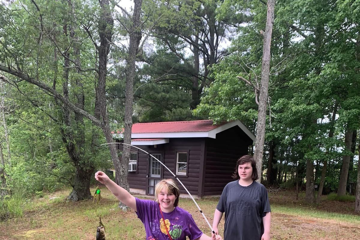 Two people standing in a grassy area with trees and a wooden cabin in the background.