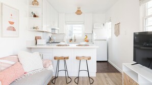 Eine moderne Küche mit weißen Schränken, einer zentralen Kochinsel mit Hockern, einem eingebauten Backofen und einem Fernseher auf einer weißen Konsole.