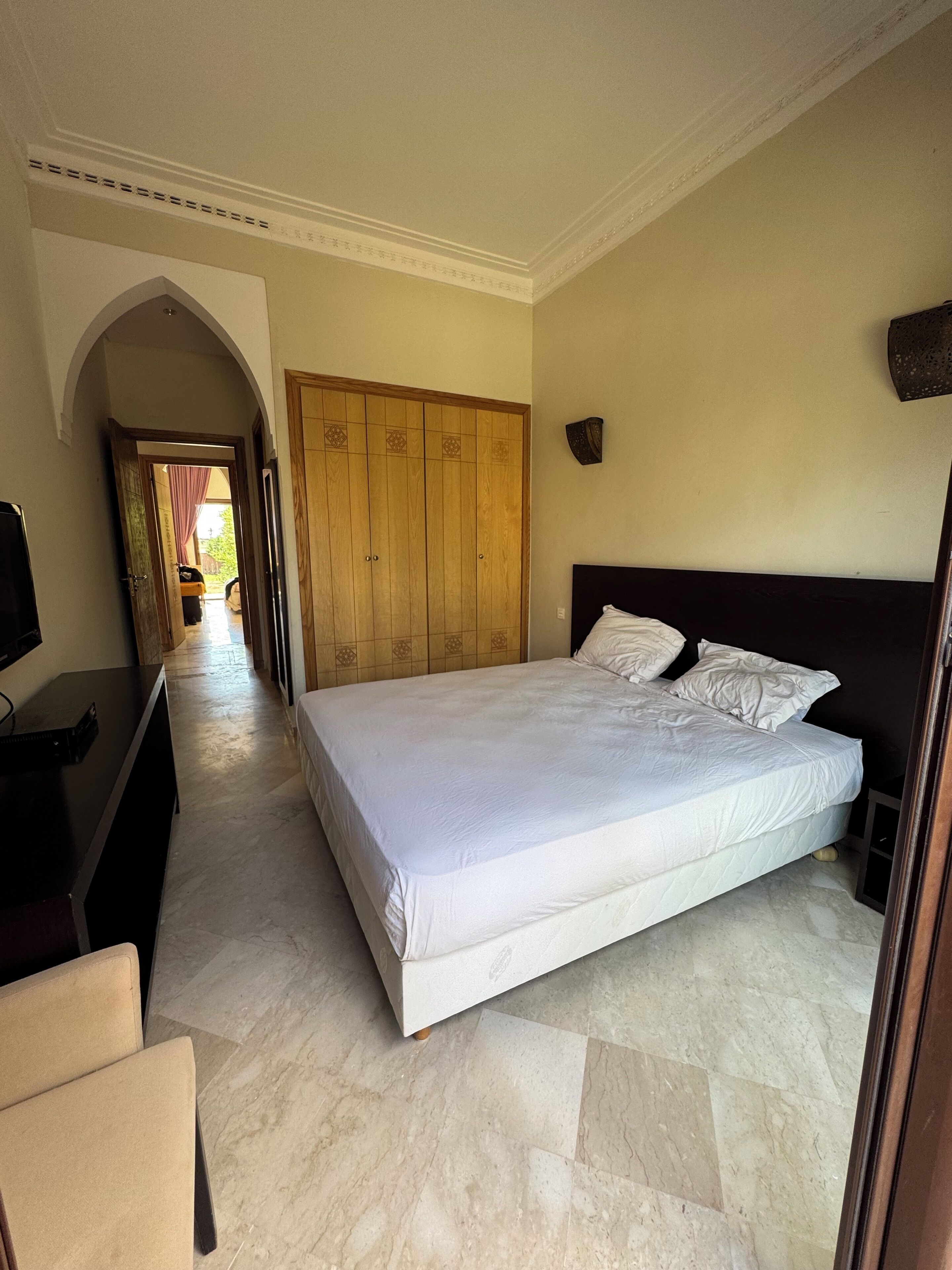 A bedroom with a large bed, a wooden wardrobe, a television, and a marble floor.