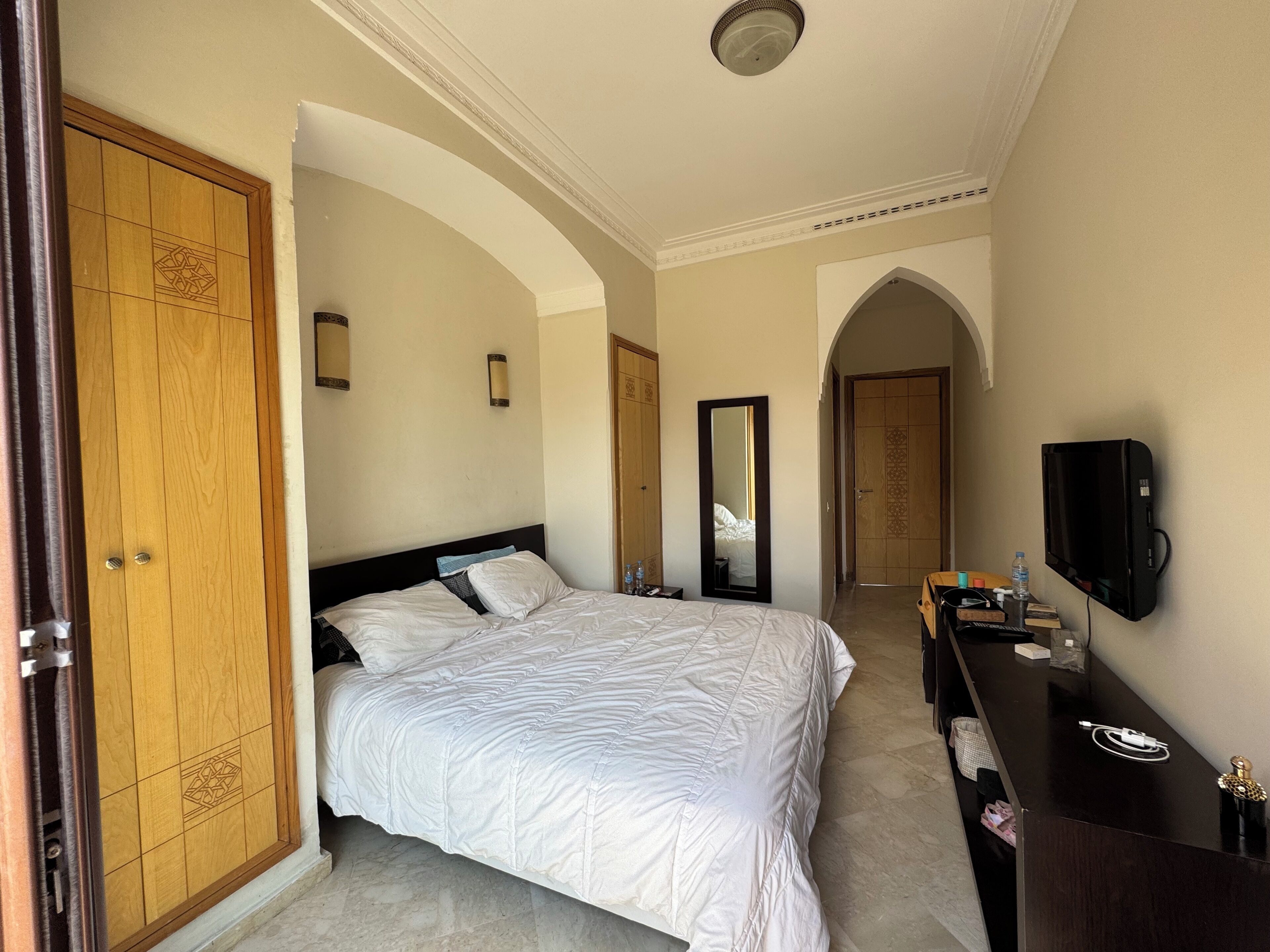 Ein Hotelzimmer mit einem großen Bett, einem Kleiderschrank, einem Spiegel, einem Fernseher und einem Schreibtisch mit verschiedenen Gegenständen.