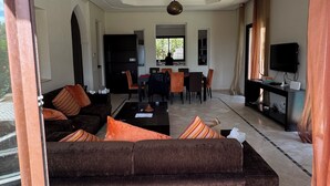 A spacious living room with a dark brown sofa, a dining area, and a TV. - Beautiful family villa near Marrakech, with large garden, private swimming pool (Centre Commune Ouled Hassoune)
