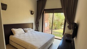 A bedroom with a bed, a desk with a computer, a window with curtains, and a view of greenery outside. - Beautiful family villa near Marrakech, with large garden, private swimming pool (Centre Commune Ouled Hassoune)