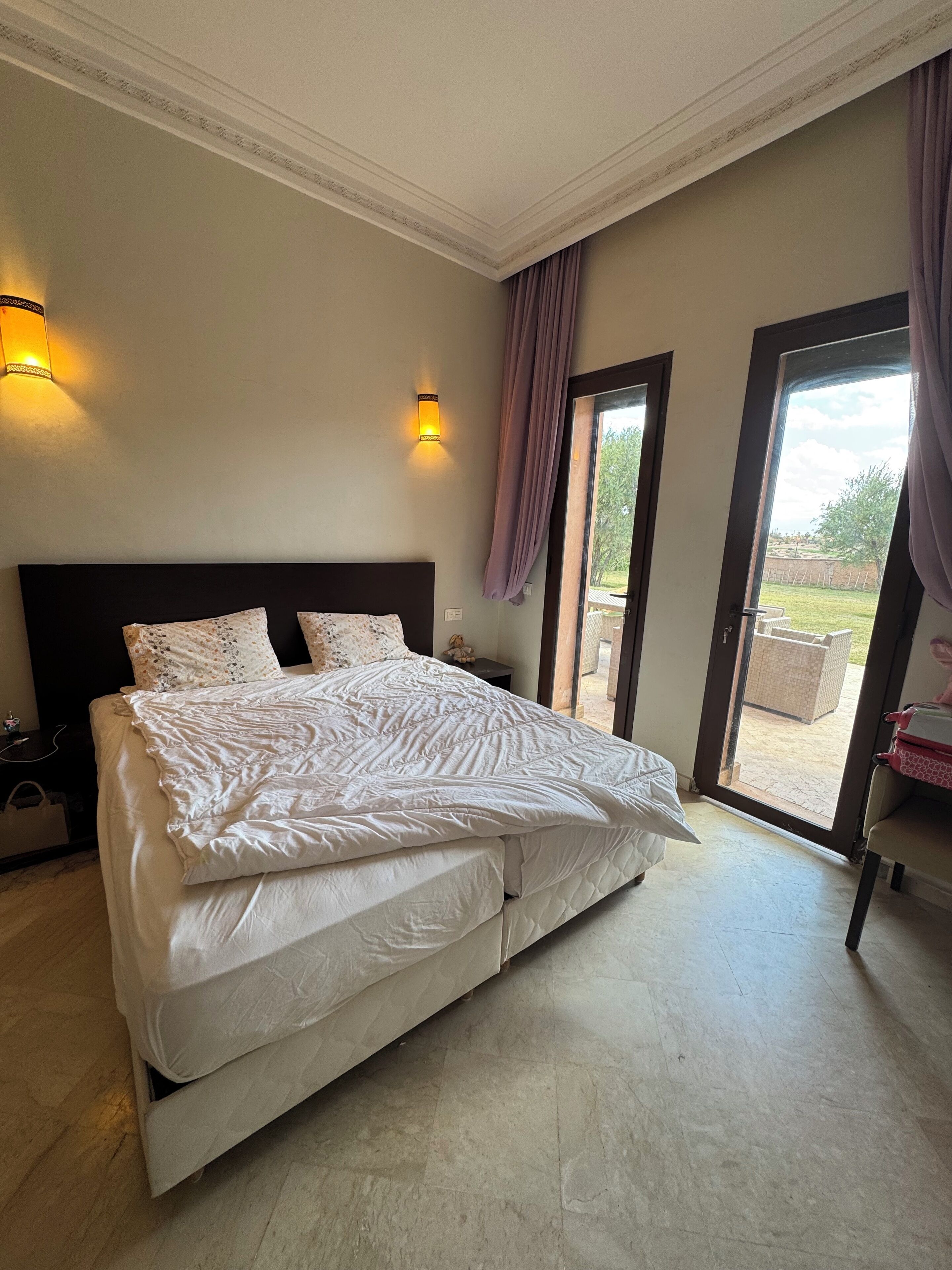 Une chambre avec un grand lit, deux appliques murales, une porte-fenêtre coulissante donnant sur l’extérieur et une table de chevet avec une lampe.