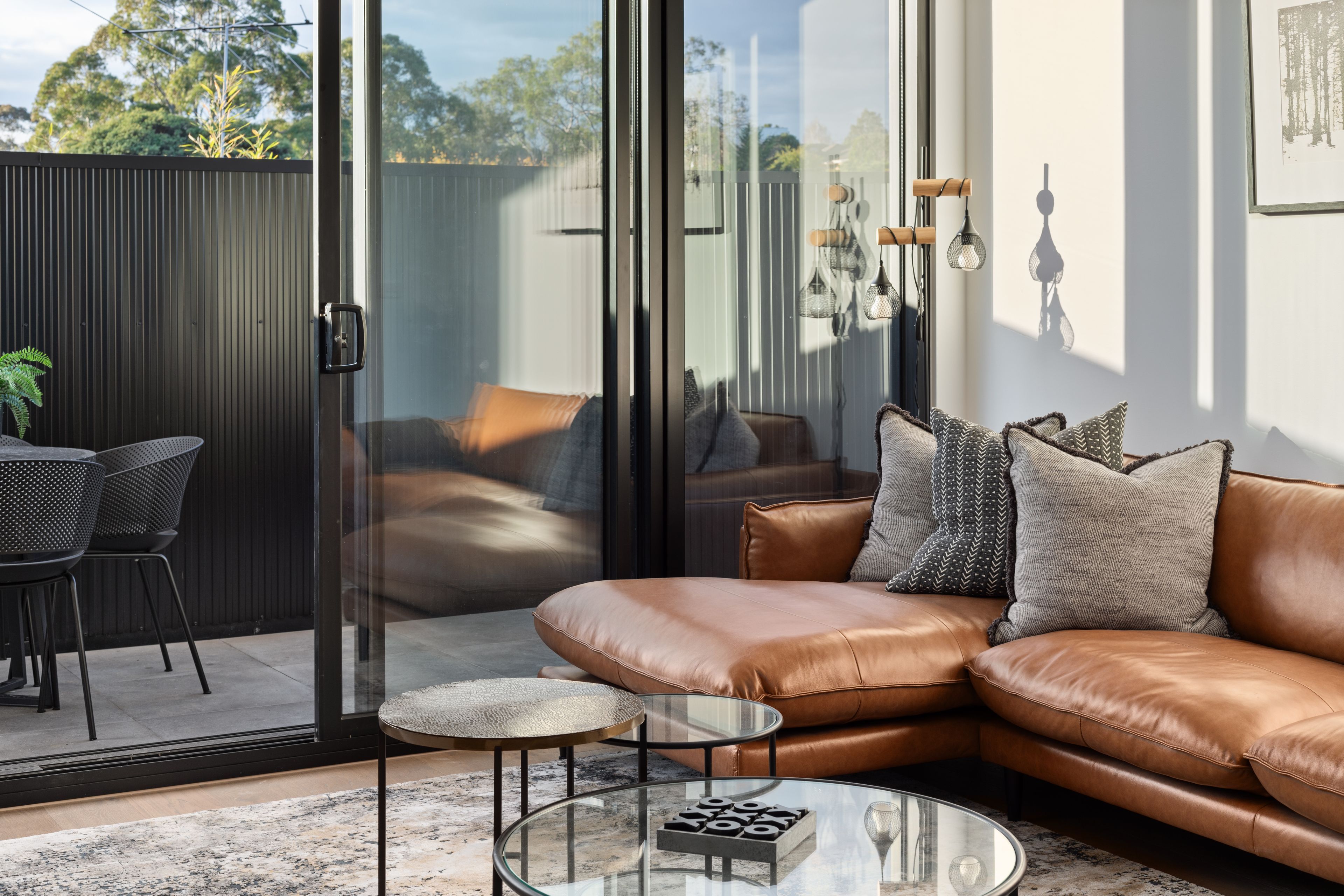 A modern living room with a leather sofa, a glass coffee table, and a view through large sliding glass doors.