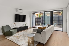 Un salon moderne avec un téléviseur à écran plat, un canapé gris, un fauteuil vert, une petite table ronde et une grande fenêtre donnant sur un jardin.