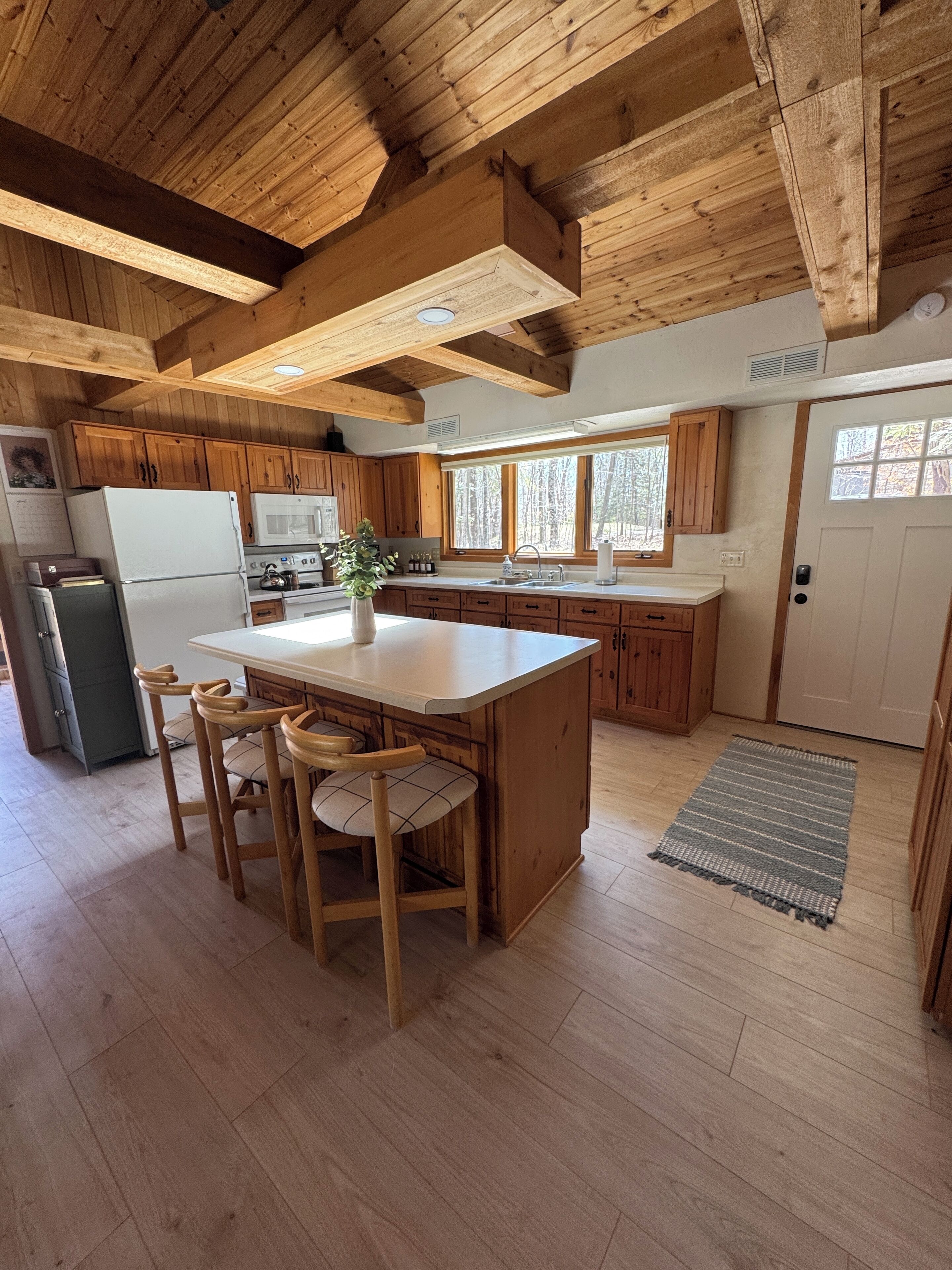 A kitchen with wooden cabinets, a central island with stools, a refrigerator, a microwave, and a window overlooking a forest.