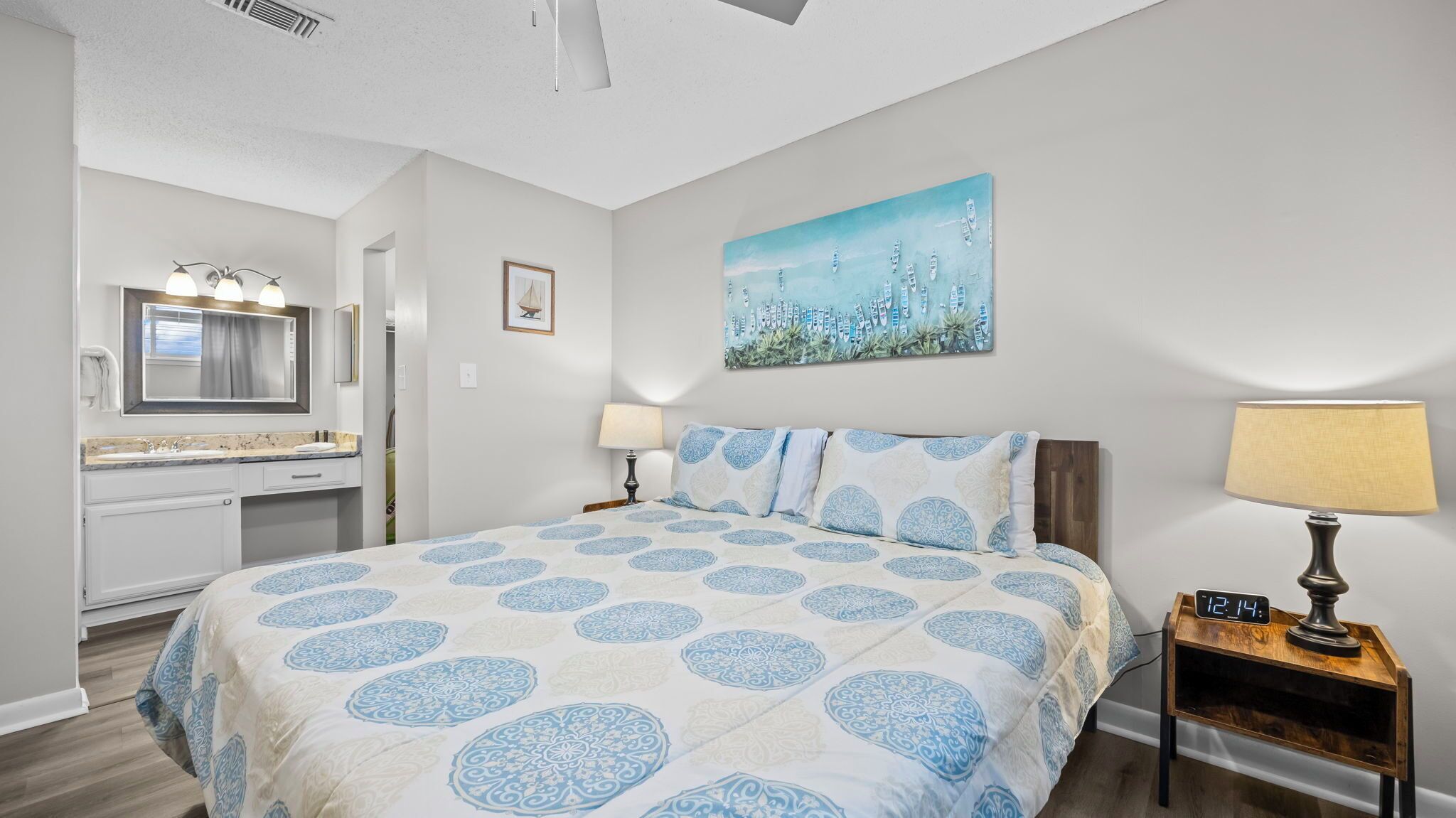 Un dormitorio con una cama grande, una mesita de noche con un reloj, un cuadro en la pared y un baño con un espejo y lavamanos.