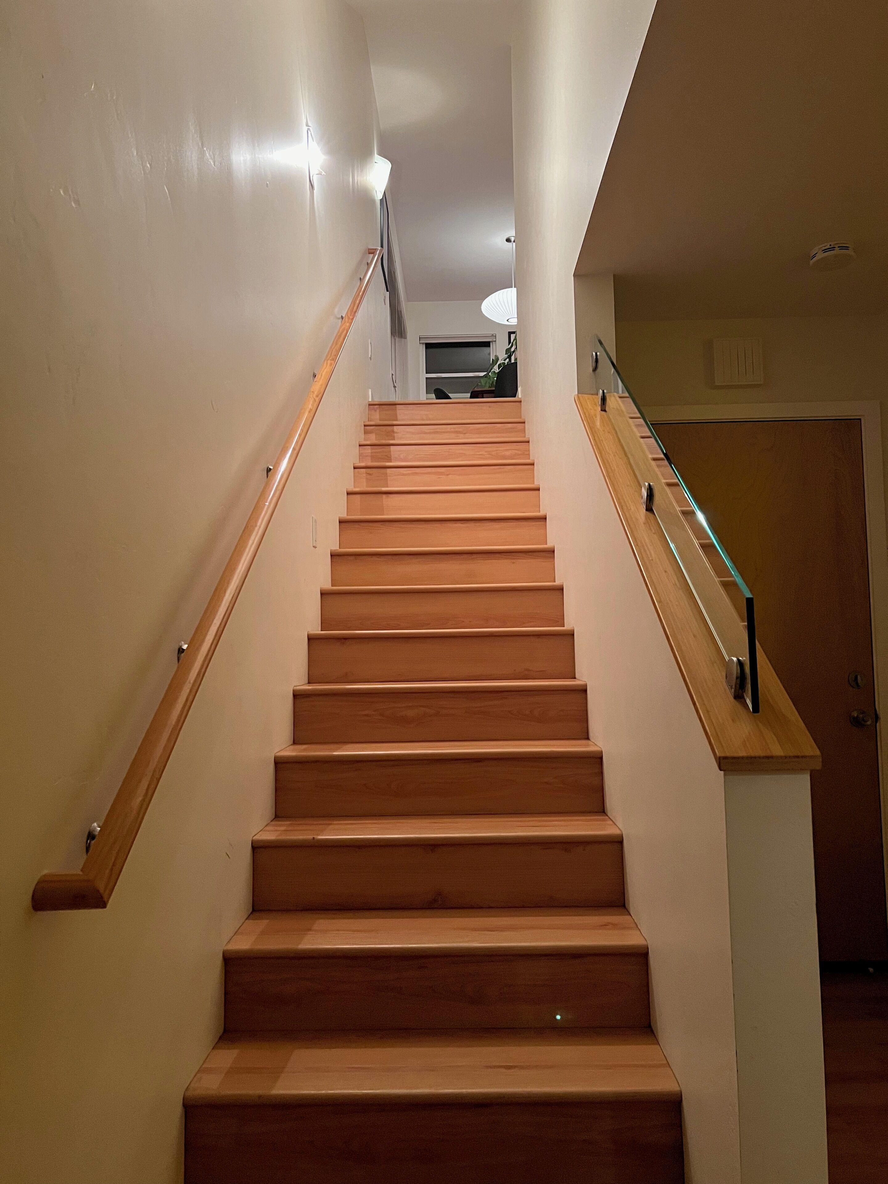 A wooden staircase with a glass handrail, leading to a landing with a door and a ceiling light fixture.