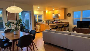 A modern kitchen with wooden cabinets, a central island, and pendant lighting. There is a dining area with a table and chairs, and a grey sofa in the living room. - Stylish, Modern 3-Level House-Yard-Patio-Balcony-Sauna- Blocks to Beaches - OB  (San Diego)