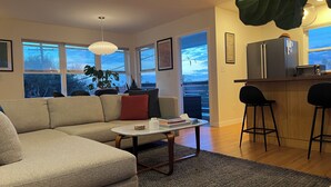 A modern living room with a sectional sofa, a coffee table, and a kitchen area with a refrigerator and bar stools. - Stylish, Modern 3-Level House-Yard-Patio-Balcony-Sauna- Blocks to Beaches - OB  (San Diego)