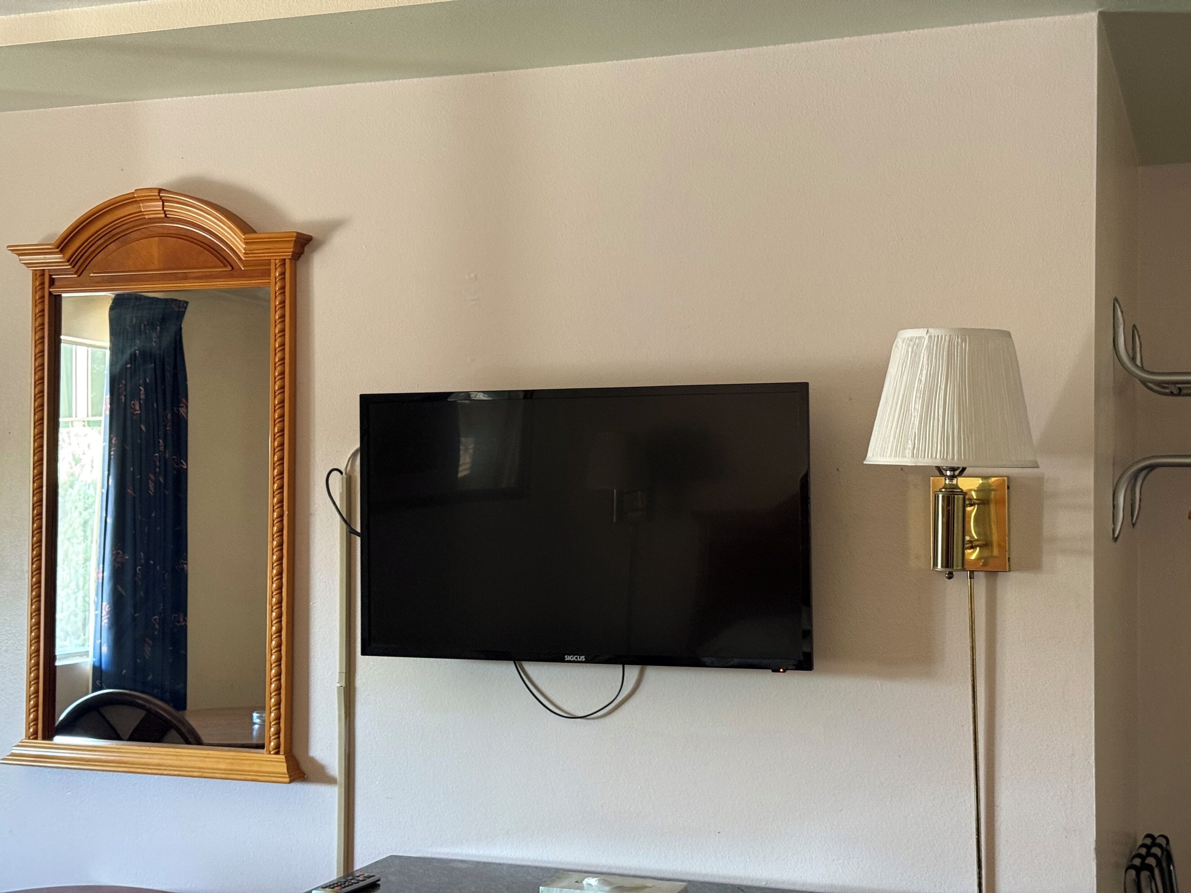 A room with a flat-screen TV mounted on the wall, a mirror with a wooden frame, and a table lamp with a white lampshade.
