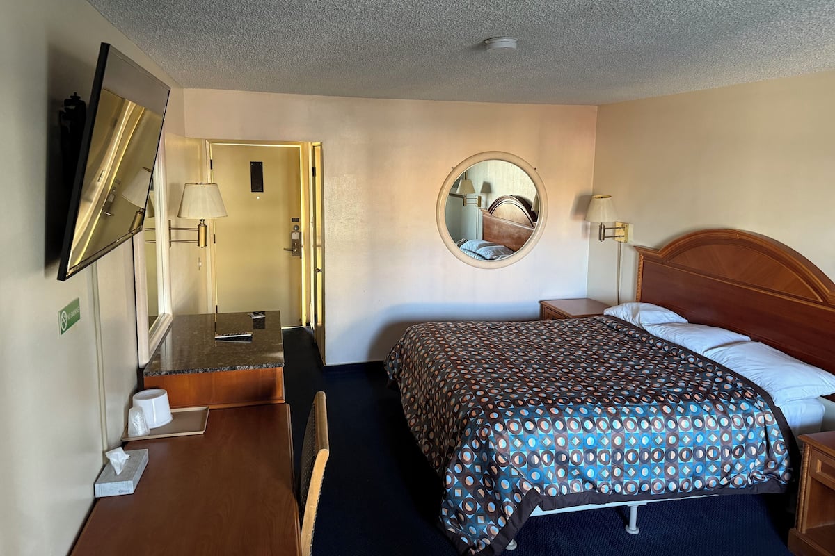 A hotel room with a bed, a desk, a chair, a round mirror, a nightstand, and a lamp.