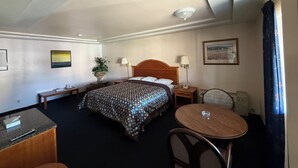 Une chambre d’hôtel comprenant un lit, des tables de chevet, un bureau, une chaise, une petite table, une lampe, une plante et un tableau au mur.
