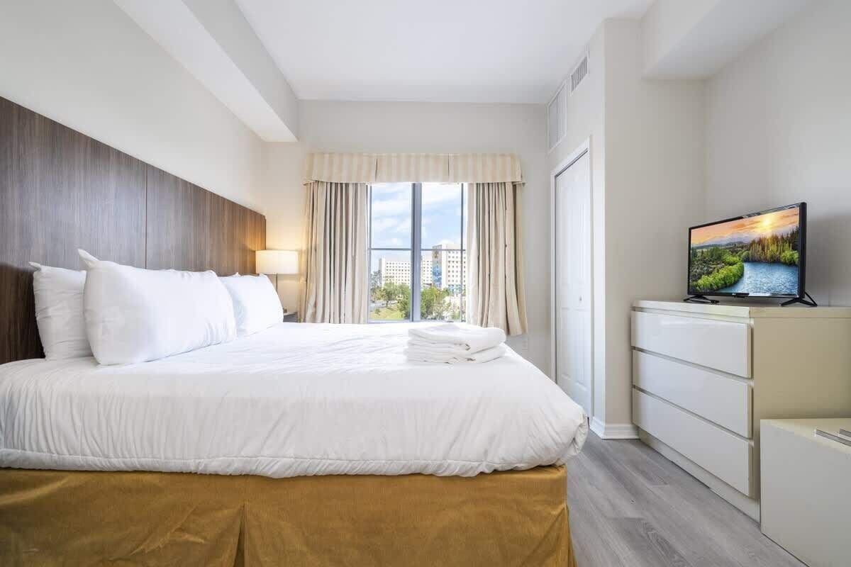Una habitación de hotel con cama, televisor, ventana con cortinas y vistas a edificios.