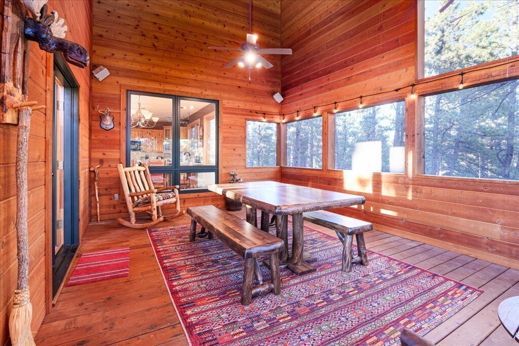L’intérieur d’un chalet en bois comprend une table à manger, des bancs, un fauteuil à bascule et une grande fenêtre donnant sur une forêt.