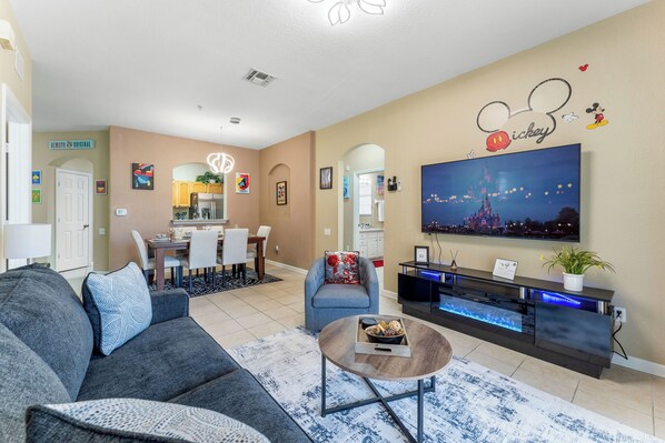 A living room with a grey sofa, a coffee table, a TV, and a wall with Mickey Mouse decorations. - Cozy 3-bedroom condo in charming Kissimmee, 2 miles from Disney world (Kissimmee)