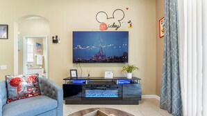 Un salon avec un téléviseur à écran plat fixé au mur, un canapé gris orné d’un coussin à l’effigie de Mickey Mouse, une table basse en bois sur laquelle sont disposés des en-cas, et un panneau décoratif sur le thème de Disney placé au-dessus du téléviseur.