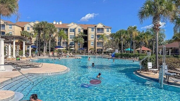 Une piscine où des gens nagent et se prélassent, entourée de palmiers et bordée d’un immeuble doté de nombreux balcons.