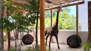 A giraffe under a wooden pergola with a gravel ground and surrounding greenery. - Tuk Tulum by Marea (Tulum)