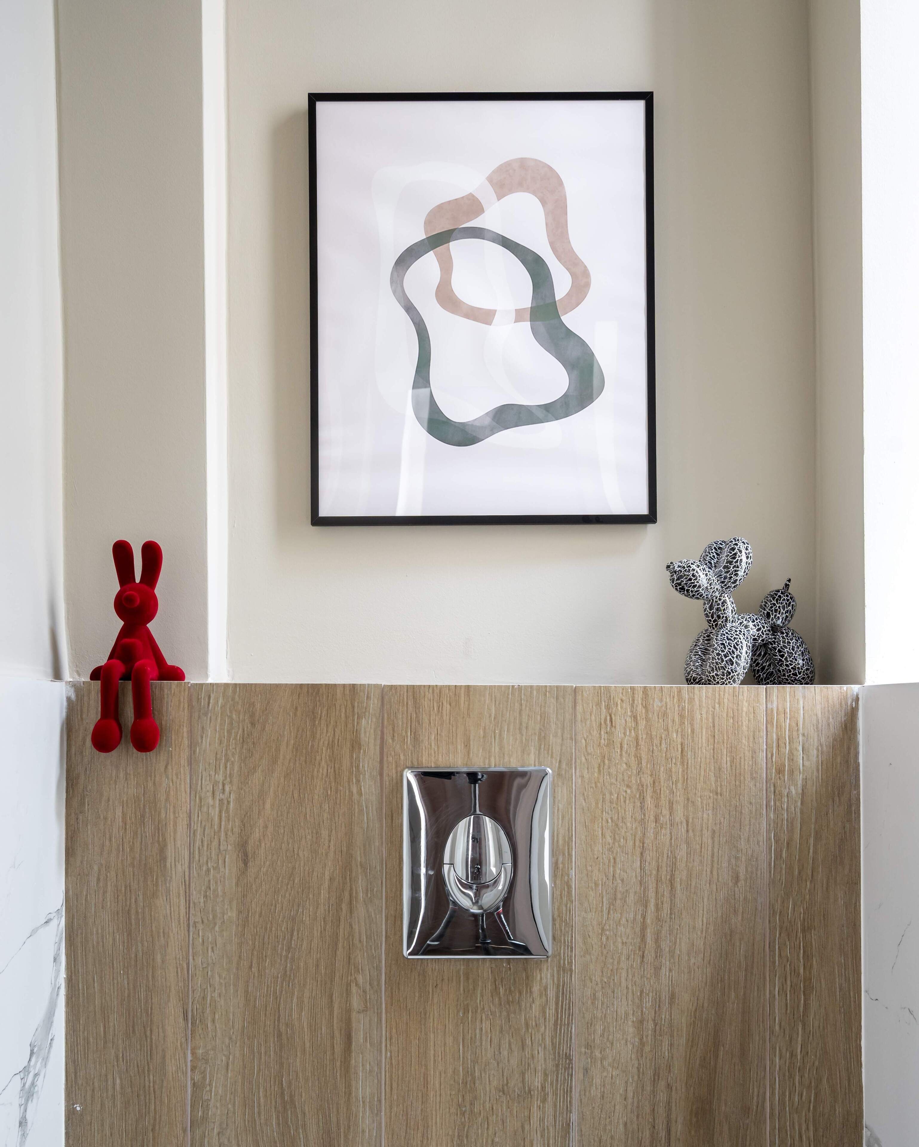 Une œuvre d’art abstraite encadrée est accrochée au mur, un jouet en peluche rouge se trouve à gauche et un objet métallique à droite.