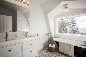 A modern bathroom with a white vanity, a large mirror, a bathtub, and a window with a view of trees and the ocean.