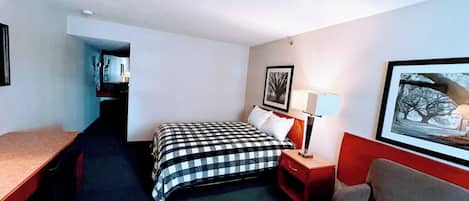 Ein Hotelzimmer mit Bett, Schreibtisch, Stuhl, Nachttisch mit Lampe und einem gerahmten Bild an der Wand.