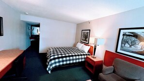 A hotel room with a bed, a desk, a chair, a nightstand with a lamp, and a framed picture on the wall.
