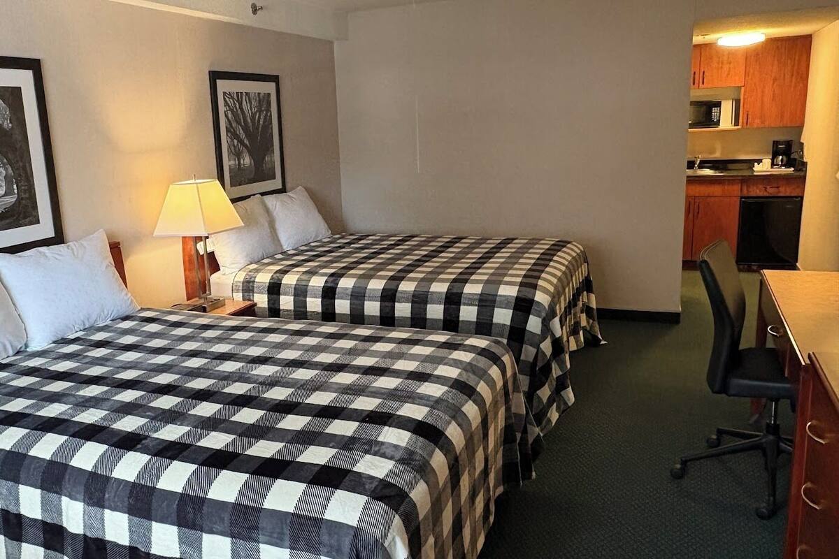 A hotel room with two beds, a desk, a chair, a lamp, and a kitchenette.
