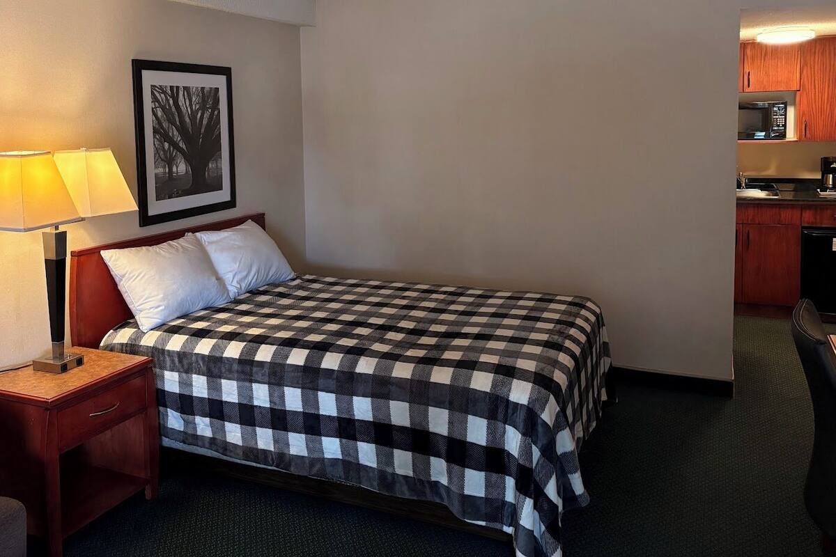 A hotel room with a bed, a nightstand with a lamp, a chair, a framed picture on the wall, and a kitchenette in the corner.