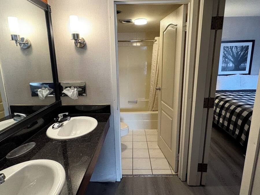 Un baño de hotel con dos lavamanos, una ducha y una cama visible a través de una puerta abierta.